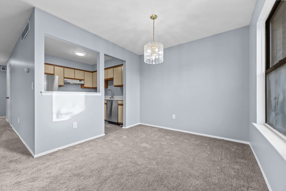 Open dining area with gray carpet, chandelier, and view into kitchen with light wood cabinets.