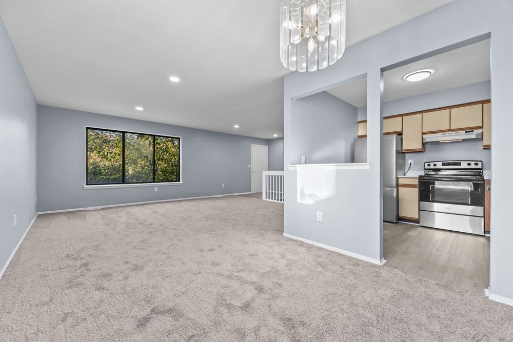 Spacious living room with gray carpet, recessed lighting, chandelier, and open kitchen view.