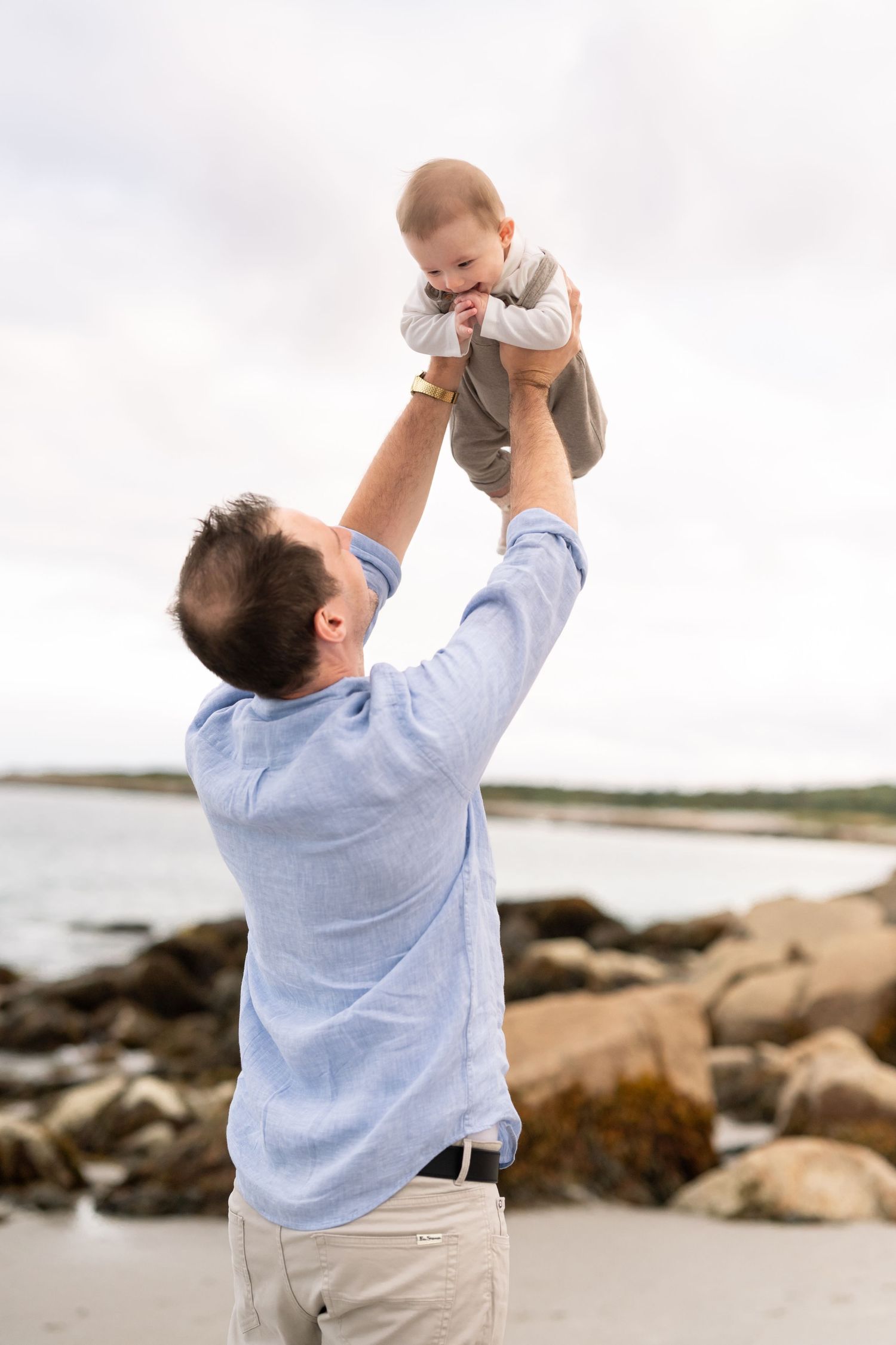 Dad holding up 3 month old baby above his head while the baby giggles during a beautiful beach photo session