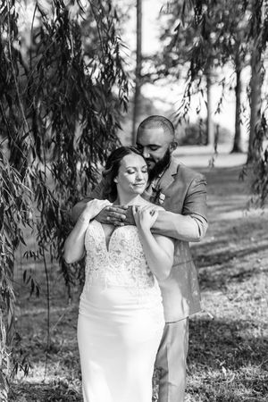 Black and white romantic portrait of groom kissing bride on shoulder under weeping willow tree.