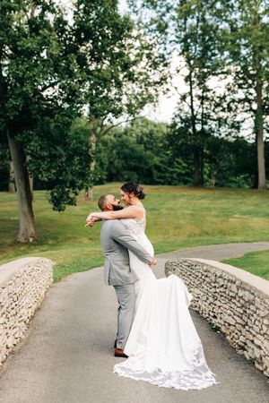 Groom lifts bride in a romantic embrace on a stone bridge surrounded by lush green parkland trees.