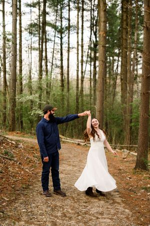 Couple dancing on a forest path, groom in navy spinning bride in flowing white dress among tall pine trees.
