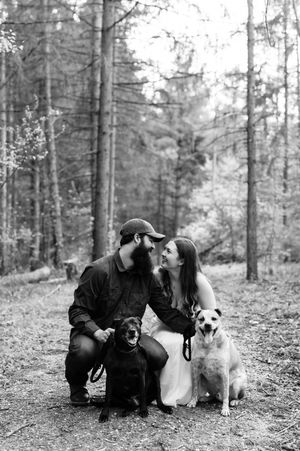 Black and white photo of a couple sharing a romantic moment with two dogs in a forest setting.