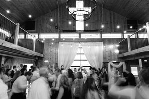 Black and white photo of a lively wedding reception in a rustic barn venue with guests dancing under warm lights.