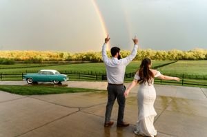 Bride and groom celebrate under a stunning double rainbow at an outdoor venue with a vintage teal car nearby.