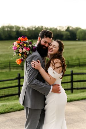 Smiling bride embracing bearded groom in gray suit holding vibrant colorful bouquet at golden hour outdoor wedding.