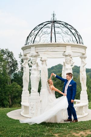 Bride and groom dancing near elegant white gazebo with ornate sculptures and iron dome on lush green hillside.