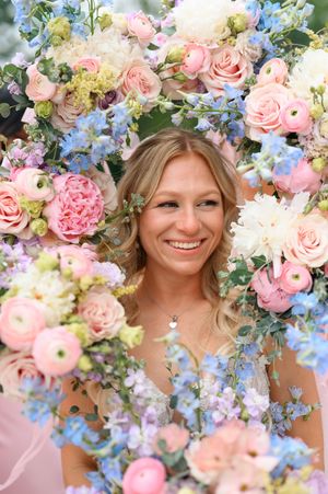 Smiling bride framed by colorful floral arrangement of pink roses, ranunculus, and blue delphinium flowers.