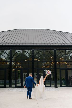 Bride raises bouquet as couple stands before glass greenhouse wedding venue with string lights.