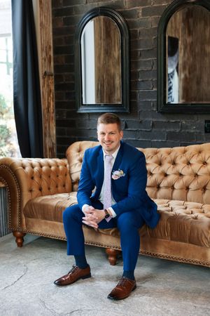 Smiling groom in blue suit sitting on tufted leather sofa with arched mirrors on brick wall behind him.
