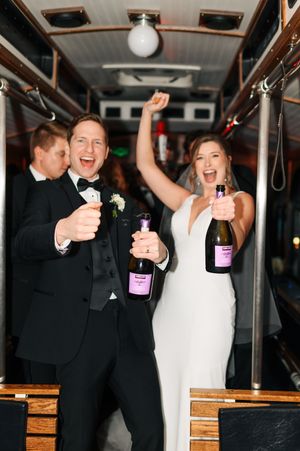 Bride and groom celebrating on party bus holding champagne bottles, dressed in wedding attire, smiling and cheering.