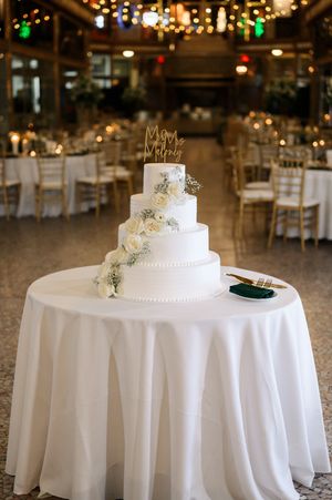 Elegant five-tier white wedding cake with roses and gold topper on draped table in warmly lit reception hall.