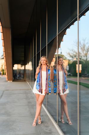 Blonde graduate in white dress with colorful stole posing beside mirrored glass building at sunset.