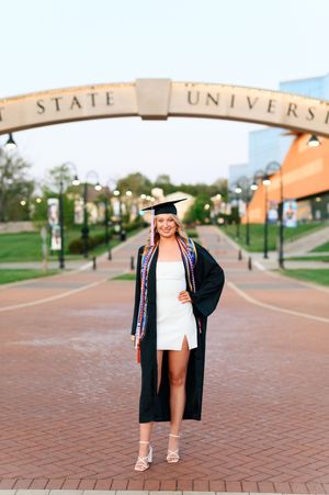 Smiling graduate in cap and gown standing under a university arch on a brick campus walkway.