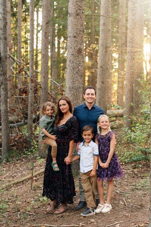 Family of five posing together in a sunlit forest with tall pine trees during golden hour.