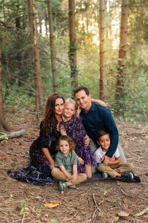 Family of five posing together in a sunlit forest, parents with three children sitting on pine needle ground.