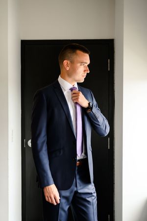 Man in navy suit adjusting purple tie while standing in doorway.