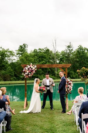 Outdoor wedding ceremony by a pond with floral arch, bride holding bouquet, groom and officiant under wooden arbor.