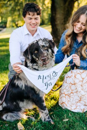 Couple smiling with their black and white dog wearing a \"She Said Yes\" engagement announcement bandana.