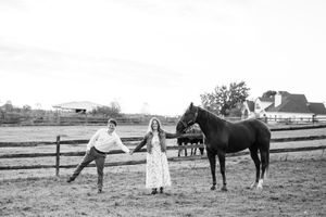 Black and white photo of a couple standing with a dark horse beside a wooden fence on a farm.