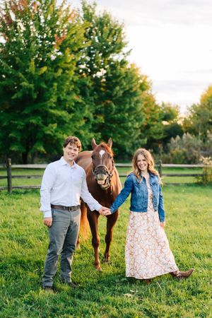 Couple holding hands beside chestnut horse with white blaze in green pasture at golden hour farm.