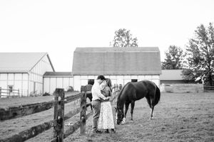 Black and white romantic couple kissing near a horse by a wooden fence at a farm.