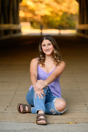 Smiling young woman in purple top and ripped jeans sitting under covered bridge in autumn.