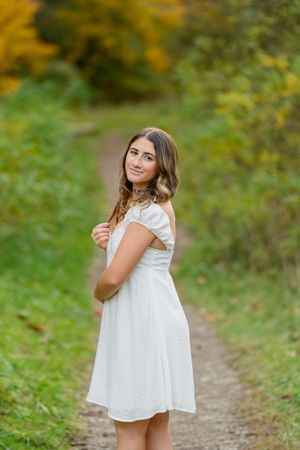 Young woman in white off-shoulder dress smiling on autumn forest path with colorful fall foliage background.