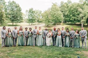 Large wedding party in sage green bridesmaid dresses and gray suits posing outdoors on lush green lawn.