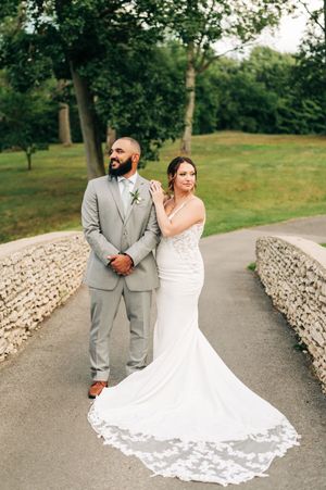 Bride in lace mermaid gown with train and groom in gray suit posing on stone bridge outdoors.