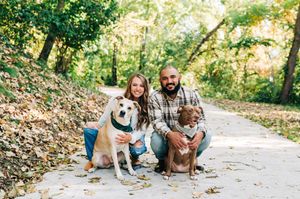 Couple crouching with two dogs on a tree-lined path covered in autumn leaves.
