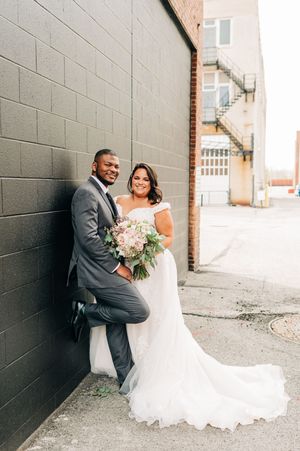 Bride and groom pose against a gray brick wall, groom in charcoal suit, bride holding a lush bouquet.