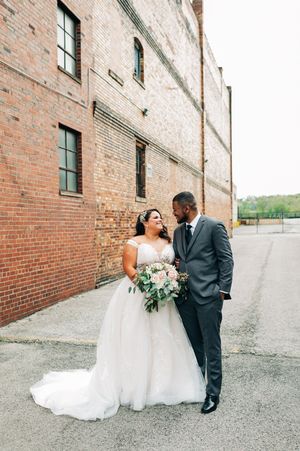 Bride and groom smiling at each other outside a brick building, bride holding a white bouquet.