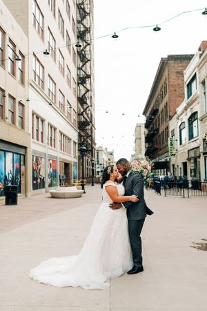 Bride and groom share a romantic kiss on a charming urban street with string lights overhead.