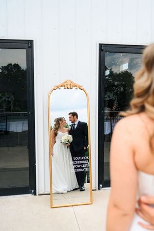 Bride and groom share a romantic moment reflected in an ornate gold mirror outside a modern white venue.