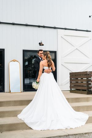 Bride and groom embracing outside a modern white barn wedding venue, bride wearing a flowing gown.