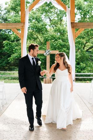 Bride in strapless gown and groom in black suit holding hands under wooden outdoor wedding ceremony arch.