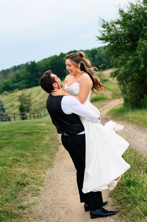 Groom lifting smiling bride in white dress on a scenic countryside path surrounded by greenery.