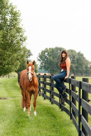 Smiling woman in casual attire sits on black farm fence beside a chestnut horse on lush green pasture.