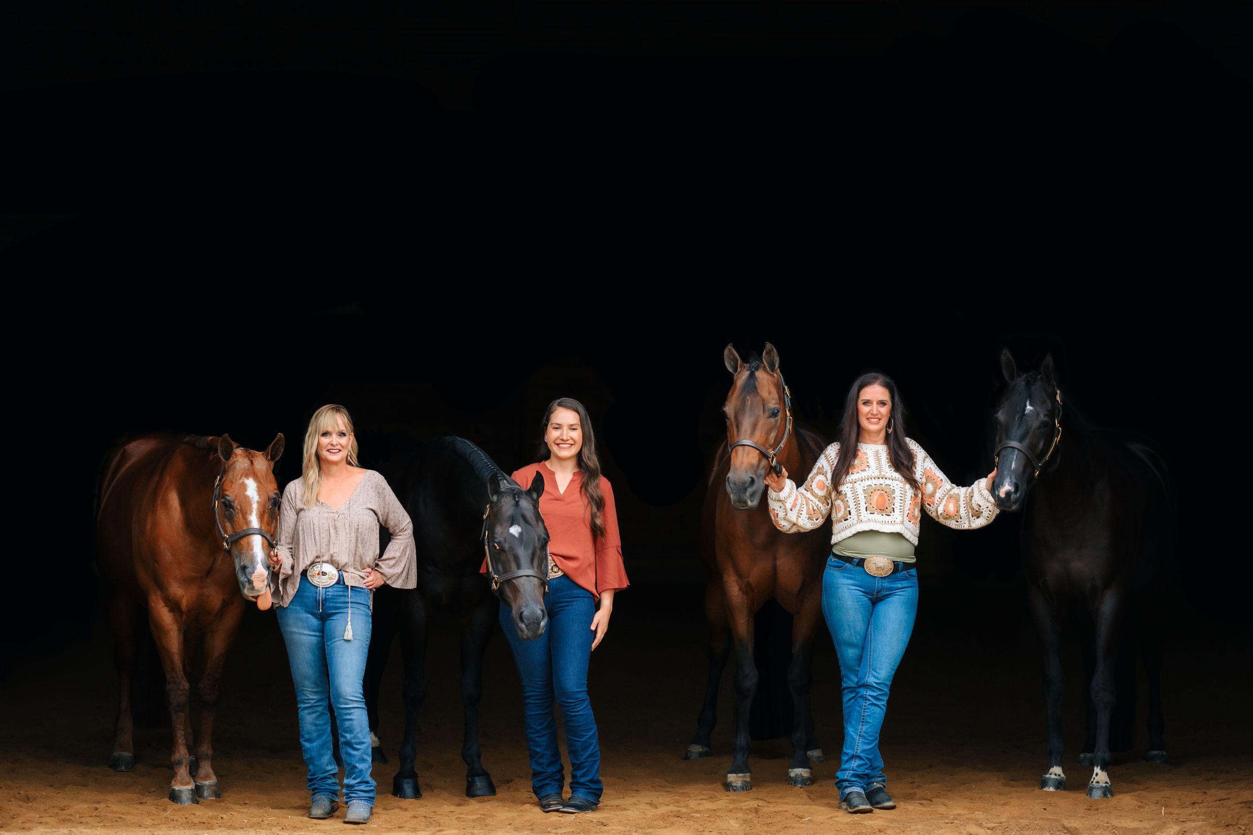 Three women in western attire posing with horses under dramatic dark arena lighting.