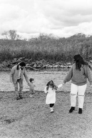 Black and white photo of a family of four holding hands near a rocky shoreline with trees in the background.