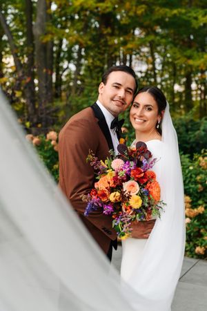 Bride and groom pose with colorful autumn bouquet outdoors, bride in white gown with veil, groom in brown suit.