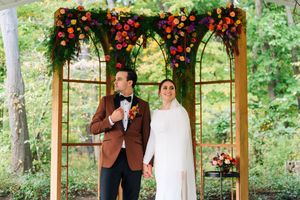 Couple stands under floral arch with vibrant red, orange, and purple blooms at outdoor wedding ceremony in wooded setting.