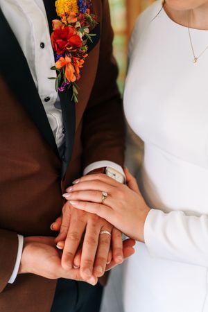 Newlyweds displaying wedding rings on joined hands, groom in brown suit with colorful boutonniere, bride in white dress.