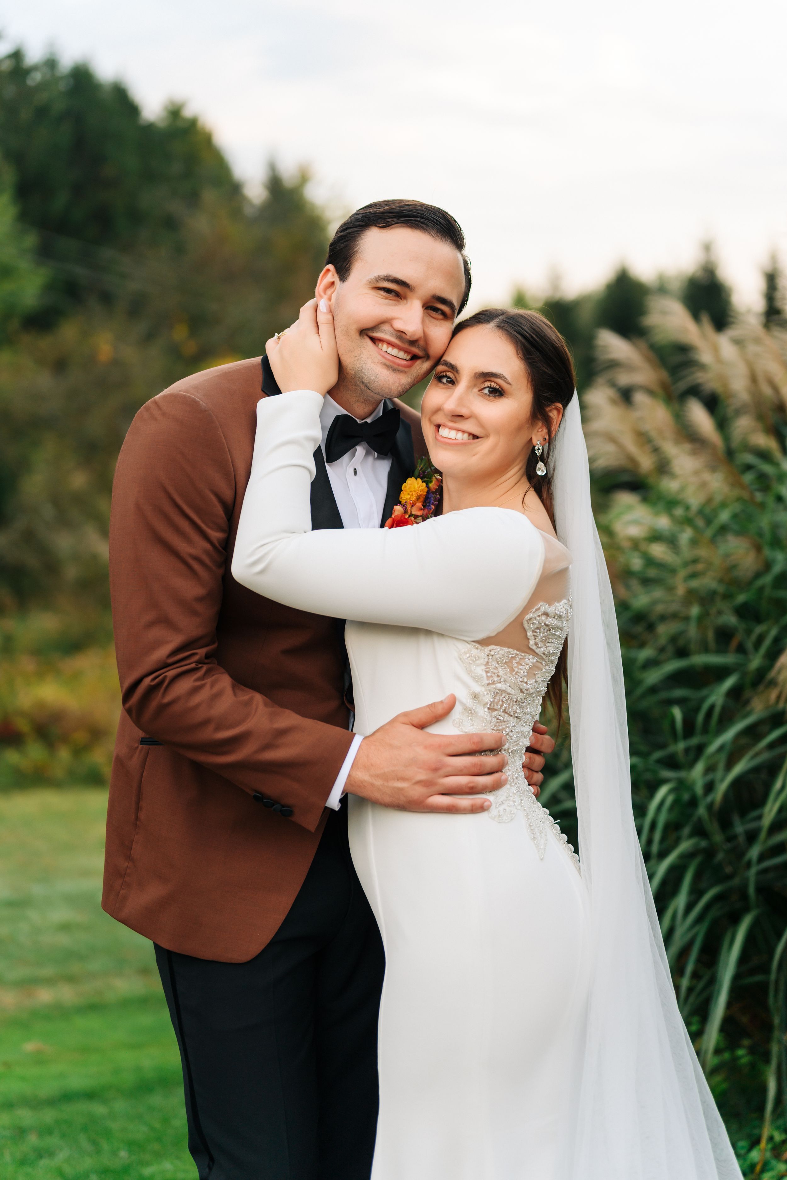 Smiling bride and groom embracing outdoors, bride in white lace gown with veil, groom in brown blazer with boutonniere.