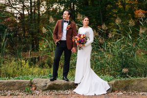 Bride in white gown holding colorful bouquet stands with groom in brown blazer on stone steps surrounded by lush greenery.