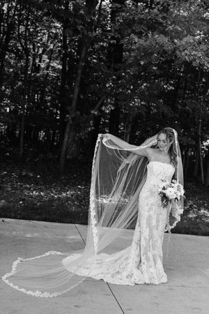 Black and white portrait of a bride in lace gown with long flowing veil billowing in the wind outdoors.