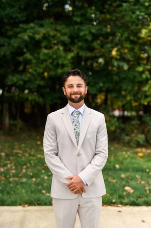 Smiling man in a light gray suit with a floral tie standing outdoors with green trees behind him.