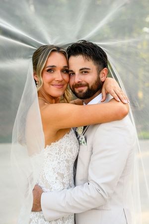 Bride and groom smiling together under a flowing wedding veil during romantic outdoor portrait session.