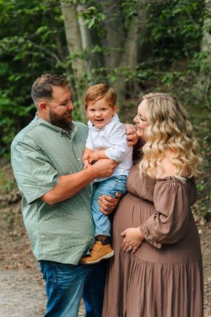Expecting family maternity photo with father holding toddler son and pregnant mother in brown dress outdoors.